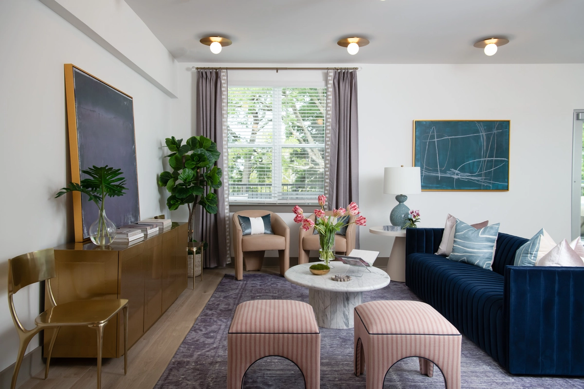 Clubhouse living area from the side showing a brass credenza with large gold-framed abstract art, fiddle leaf fig, tan barrel chairs, navy sofa with accent pillows, marble coffee table, and blush striped ottomans on a lavender area rug