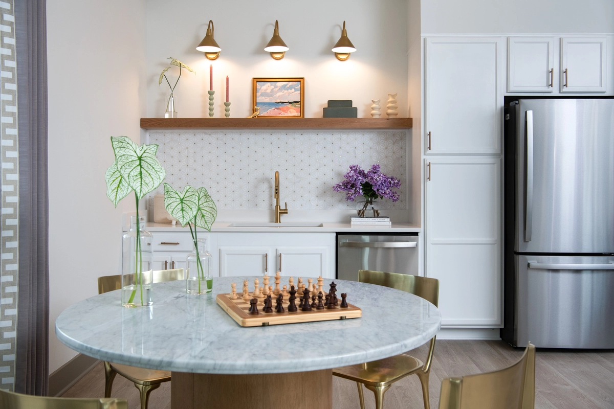 Community kitchenette dining area with round marble table, brass chairs, chess set, white cabinetry, geometric tile backsplash, brass sconce lighting, floating wood shelf with styled decor, and fresh lilacs