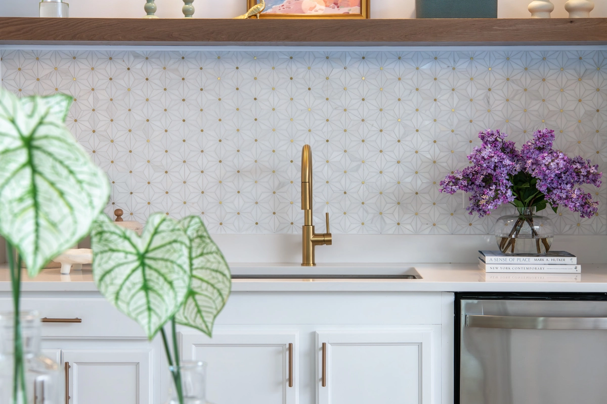 Close-up of kitchenette backsplash detail with white star-pattern mosaic tile, brass faucet, white cabinetry, floating wood shelf with decorative objects, fresh lilacs, and design books