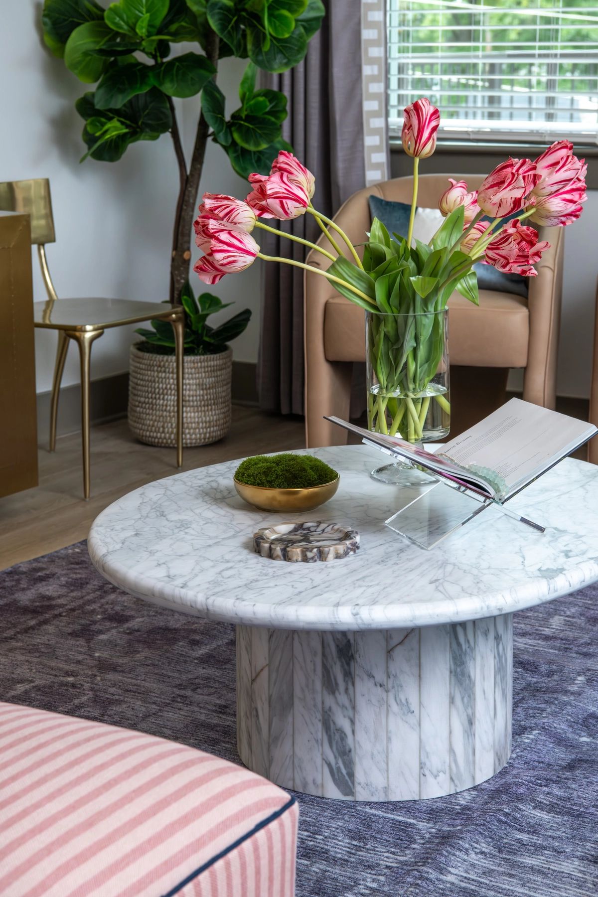Coffee table detail with white marble pedestal base, pink-and-white parrot tulips in a glass vase, open book, moss bowl, and geode accent on a lavender rug with blush ottoman and brass chair beyond