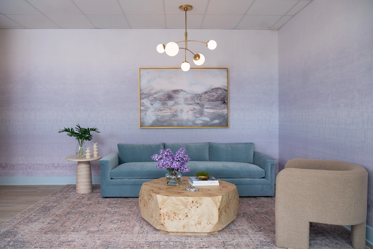 Lobby lounge with soft blue velvet sofa, burl wood geometric coffee table, brass globe chandelier, gold-framed abstract landscape painting, cream boucle accent chair, and lavender ombre wallpaper on a vintage-style area rug