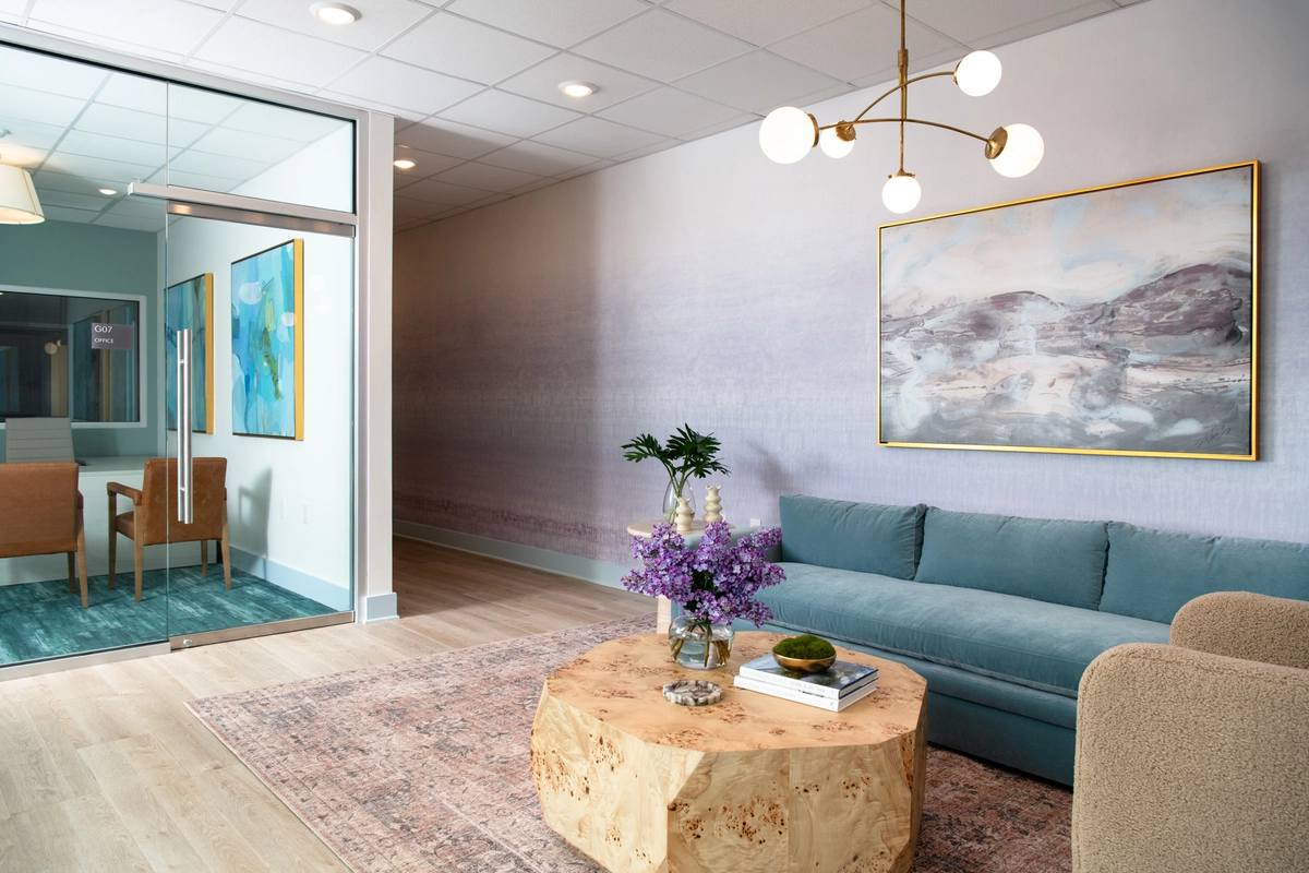 Wide view of the lobby lounge showing blue velvet sofa, burl wood coffee table with fresh lilacs, cream boucle chair, brass chandelier, lavender ombre wallpaper, and a glass-walled leasing office with teal rug and abstract art