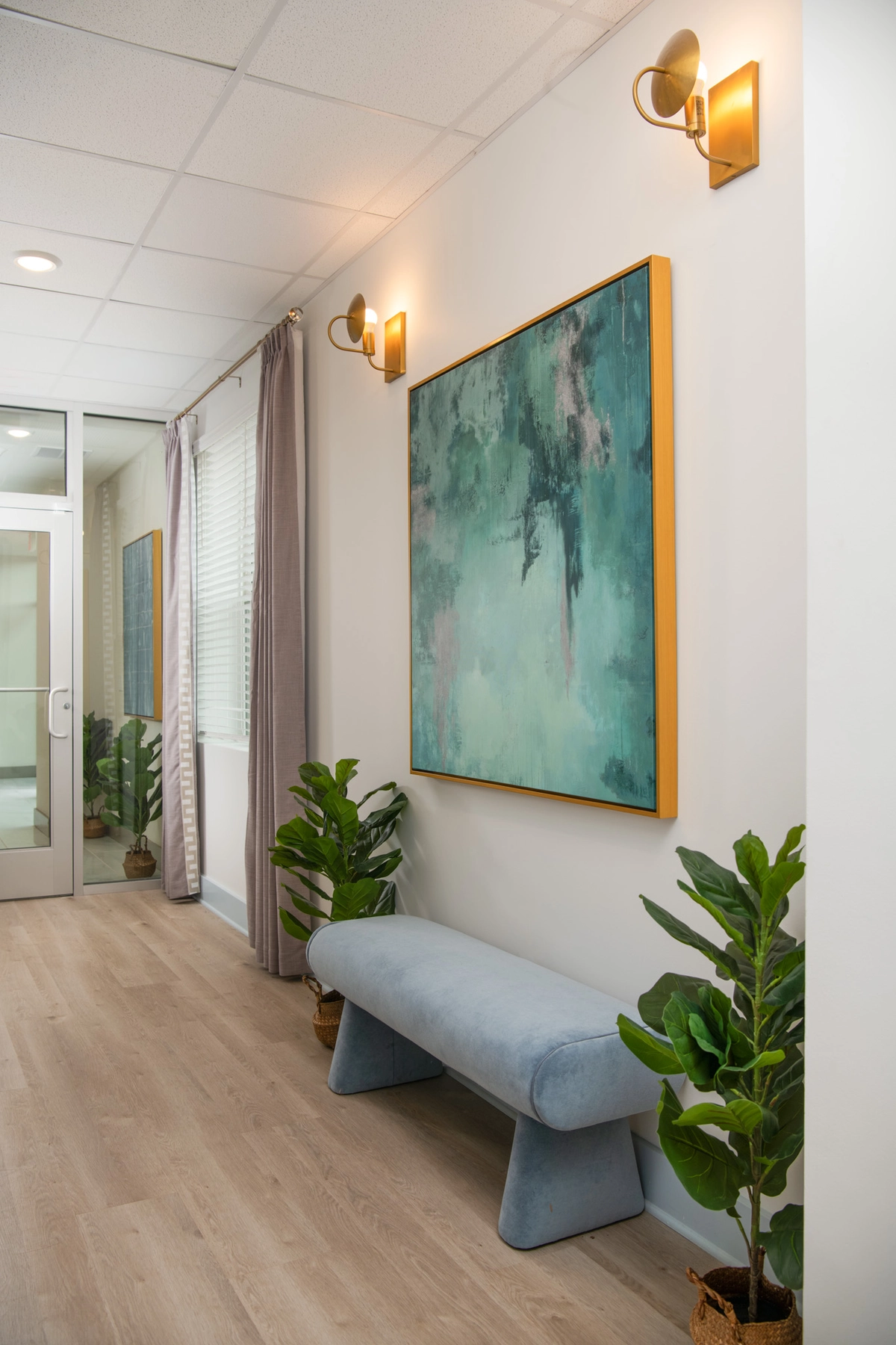 Hallway with a light blue upholstered bench, large gold-framed teal abstract painting, brass wall sconces, fiddle leaf fig plants in woven baskets, and lavender curtains on light hardwood flooring