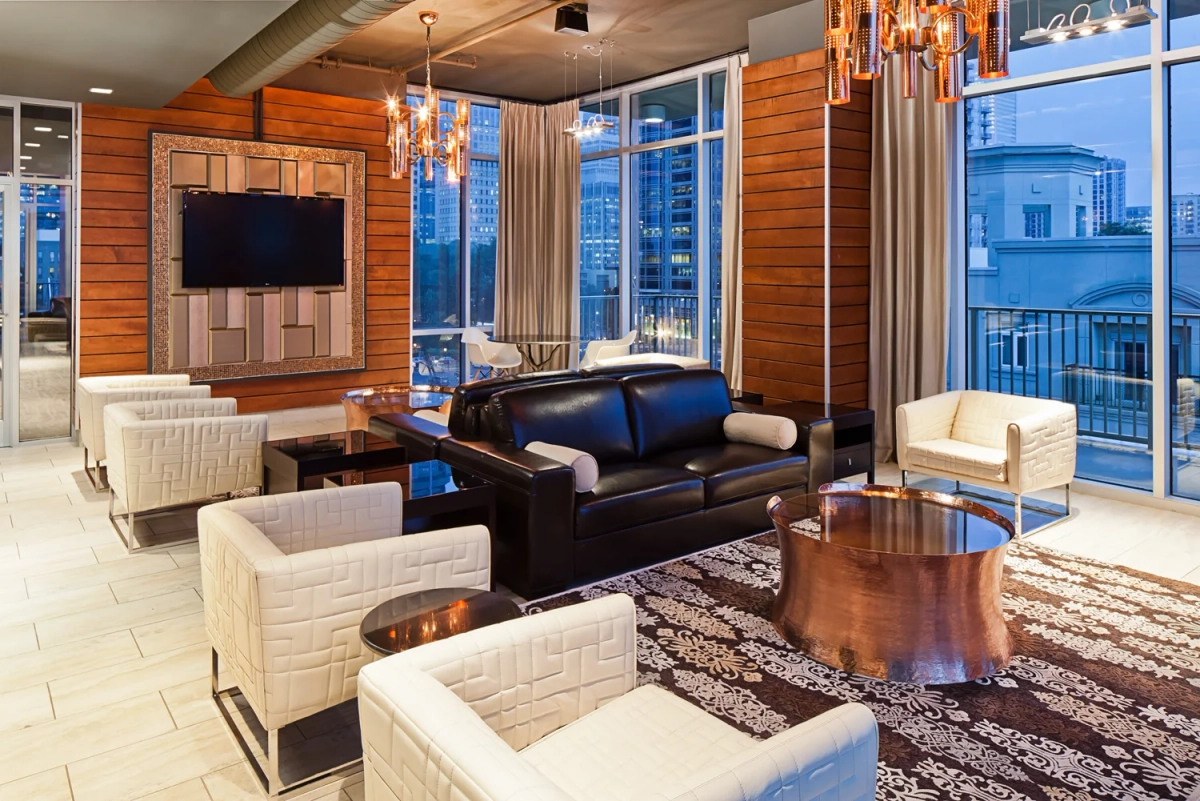 Upscale clubhouse lounge with a black leather sofa, white tufted armchairs, copper pendant chandeliers, a wall-mounted TV on horizontal wood paneling, and floor-to-ceiling windows with city views