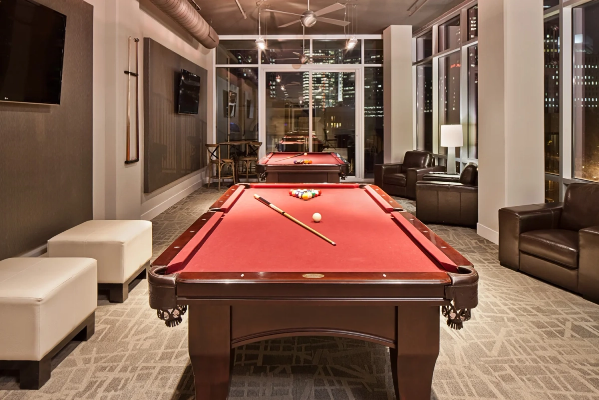 Game room with two red-felted billiard tables, dark leather lounge chairs, a wall-mounted TV, industrial-style ceiling, and floor-to-ceiling windows with nighttime city views