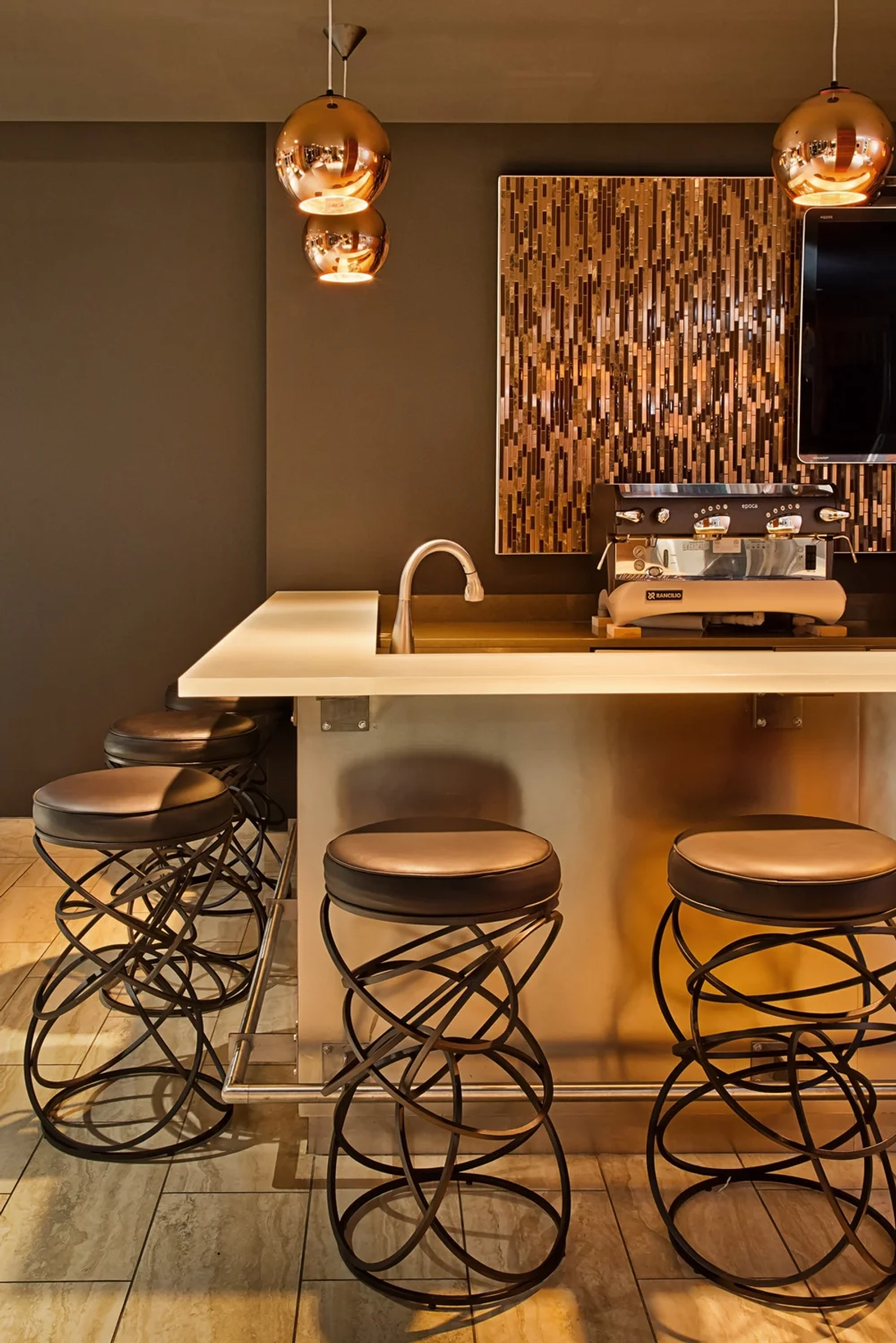 Clubhouse bar area with sculptural wrought-iron base bar stools with leather seats, copper globe pendant lights, a quartz countertop bar with sink, and a mosaic tile accent wall