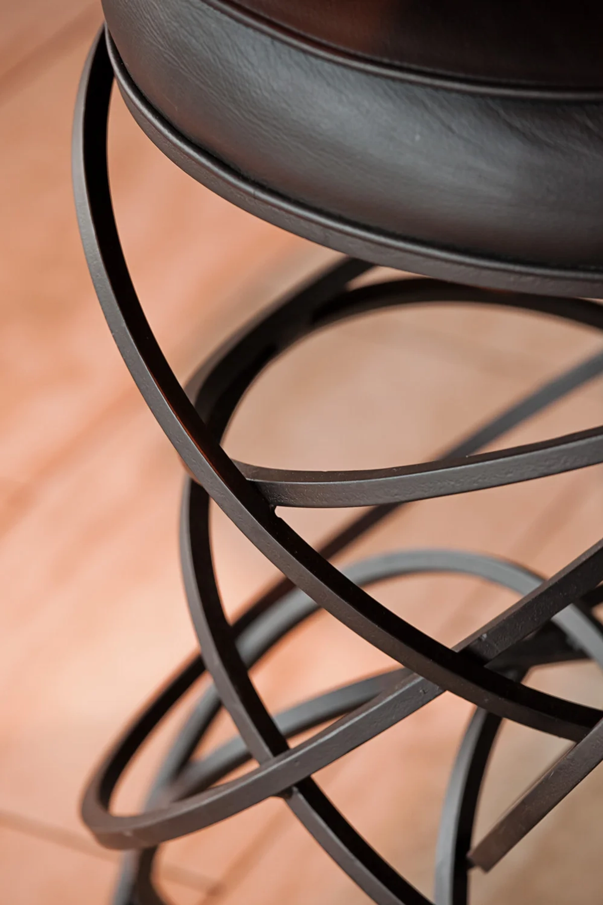 Close-up detail of a sculptural bar stool base with interlocking curved wrought-iron rings and a leather cushion seat on warm hardwood flooring