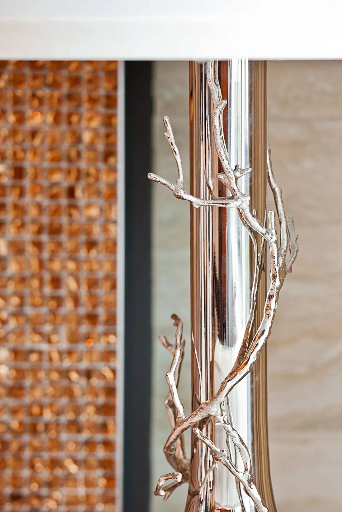 Decorative detail of a silver branch-motif sculpture wrapped around a polished chrome column, with copper mosaic tile visible in the background