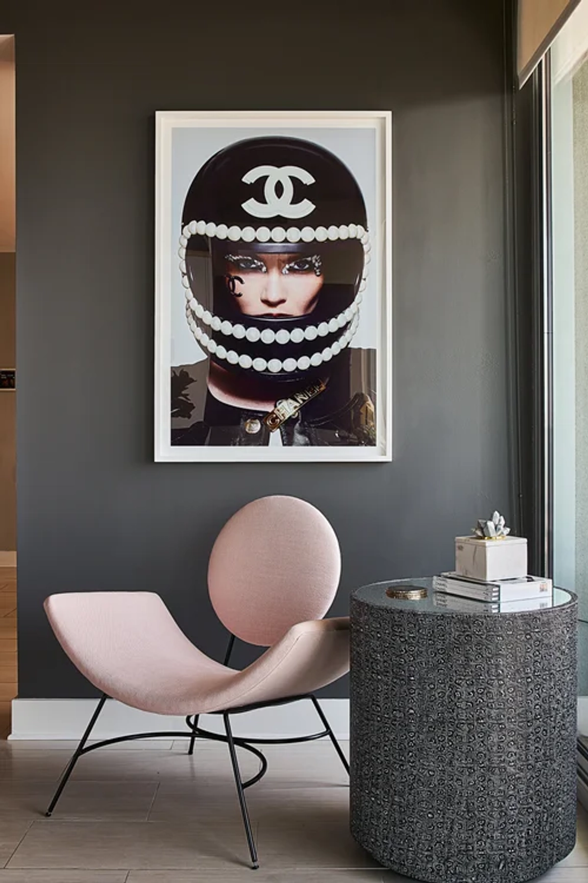 Moody vignette with a blush pink modern accent chair and a concrete side table against a dark charcoal wall, beneath a large fashion-inspired portrait with a Chanel motif