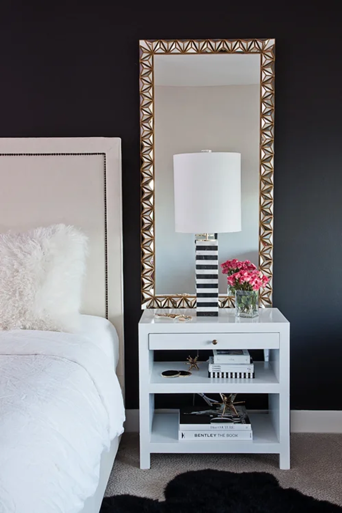 Bedroom nightstand detail with a white lacquer nightstand, a black-and-white striped table lamp, a gold geometric-framed mirror, pink roses, and a dark navy accent wall