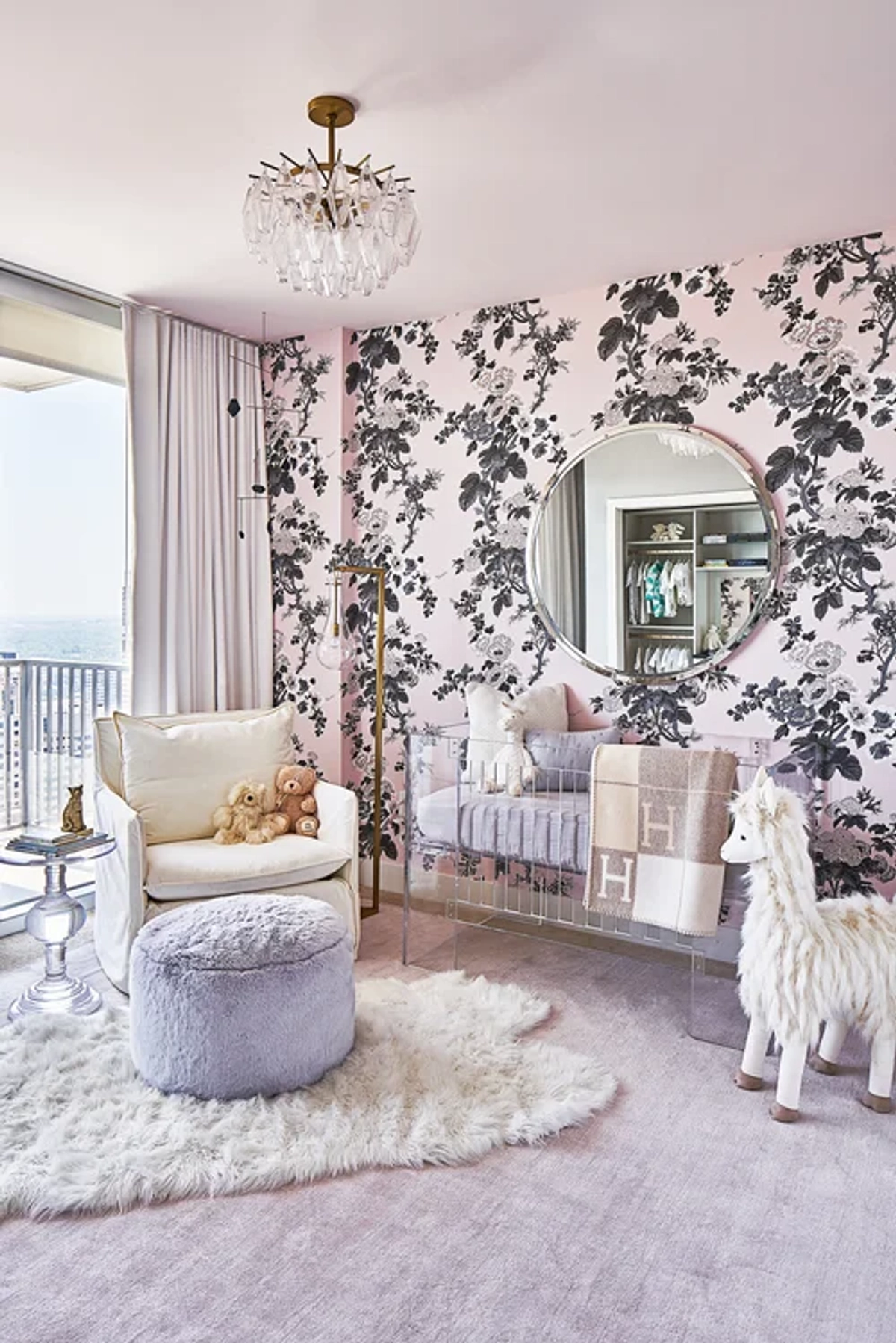 Feminine nursery with bold black-and-pink floral wallpaper, a white crib with ruffled skirt, a lavender velvet swivel chair, a crystal chandelier, and a plush white rug