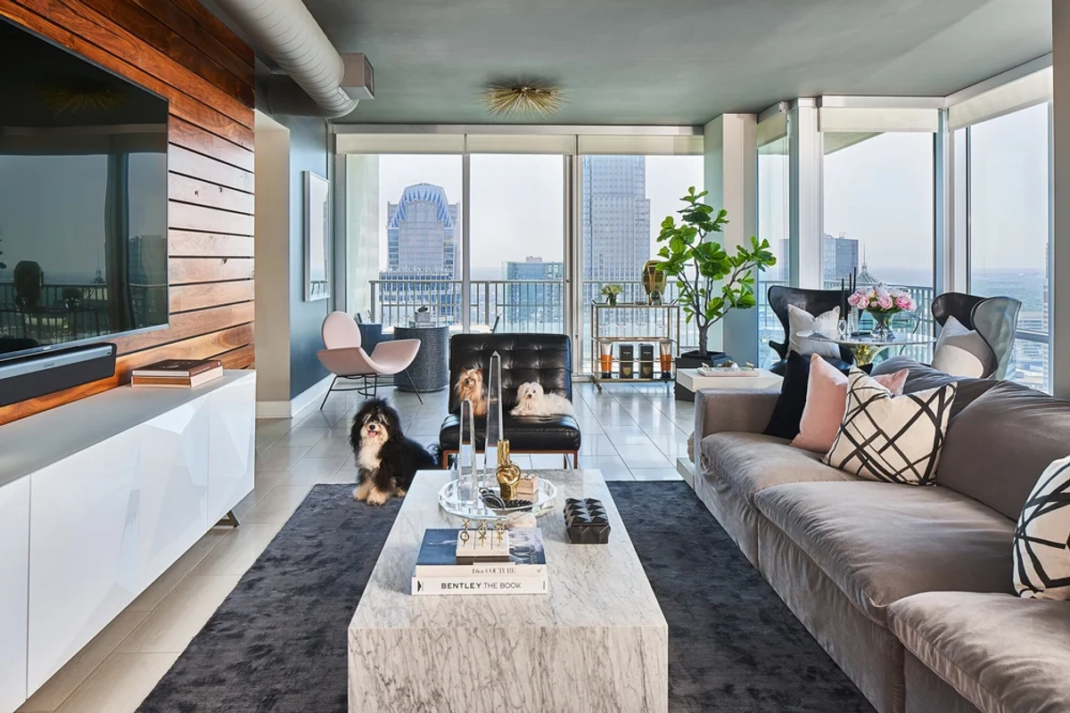 Penthouse living room with a taupe velvet sofa, a marble coffee table, reclaimed wood media wall, pink accent chairs, and floor-to-ceiling windows with Charlotte skyline views