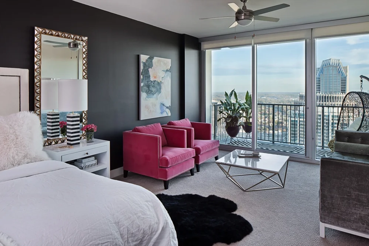 Master bedroom with a dark navy accent wall, white upholstered bed, two hot pink velvet armchairs, a chrome coffee table, abstract artwork, and floor-to-ceiling windows with city views