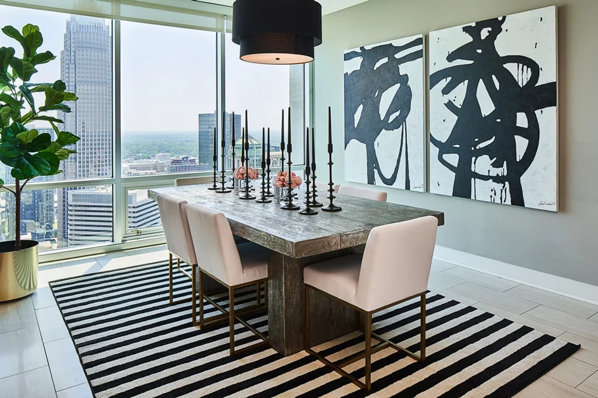 Modern dining room with a reclaimed wood table, blush upholstered chairs with gold legs, tall black candlesticks, a black drum pendant, bold abstract art, and Charlotte skyline views