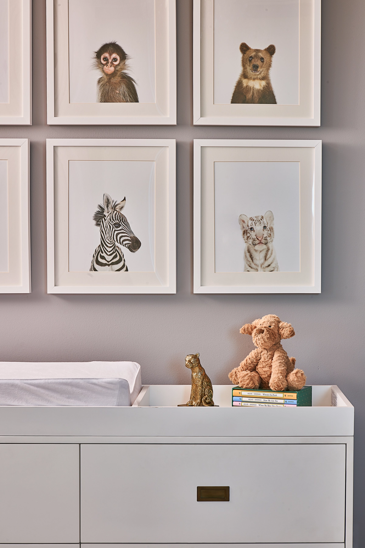 Nursery detail with a grid of four framed baby animal portraits, a white dresser with brass hardware, a gold cheetah figurine, teddy bear, and stacked children's books