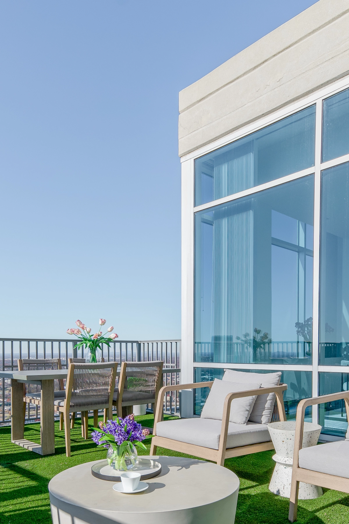 Rooftop balcony with a gray teak dining table and chairs, a cream cushioned lounge chair, white side table with purple flowers, turf flooring, and panoramic city views