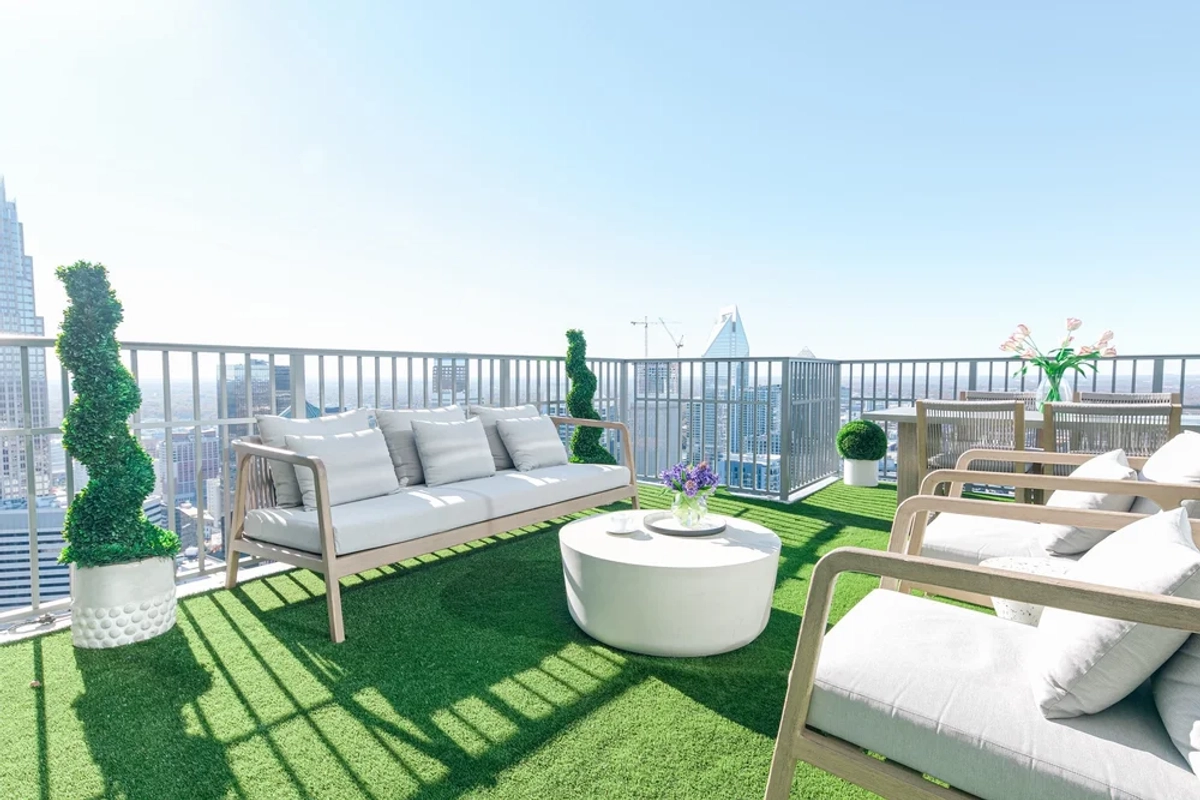 Expansive high-rise balcony with turf flooring, a white modular outdoor sofa, a round white coffee table, woven lounge chairs, and panoramic views of the Charlotte skyline