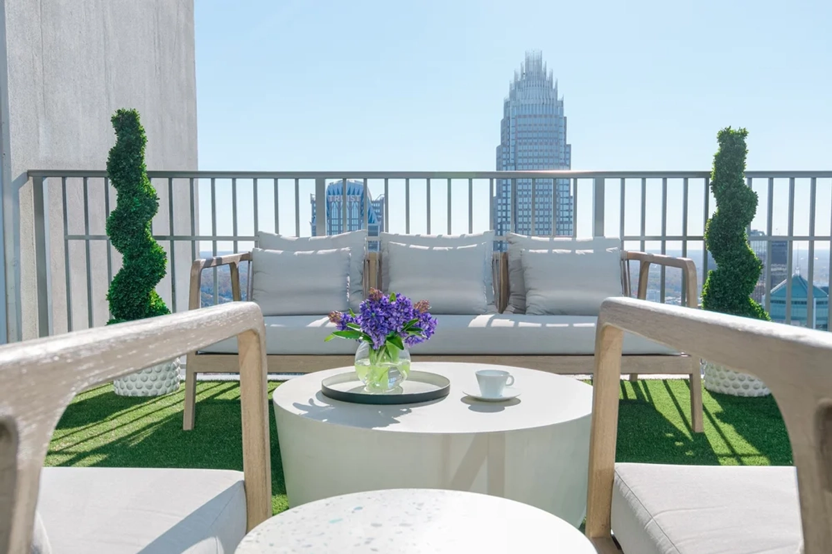 Intimate balcony dining area with a white bistro table, white woven chairs, green topiaries, a vase of purple flowers, and a close-up view of Uptown Charlotte
