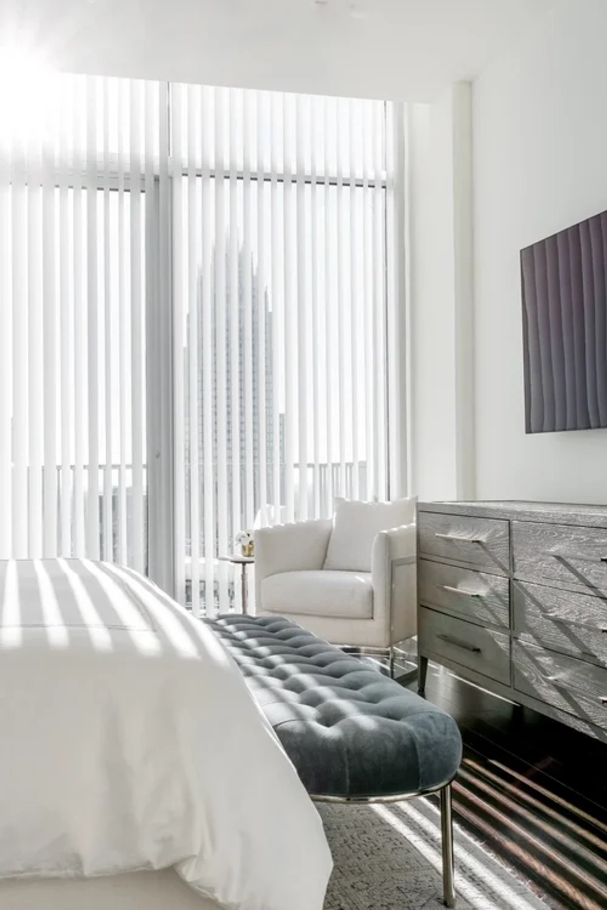 Minimalist high-rise bedroom sitting area with a white accent chair, a mirrored dresser, a tufted dark teal bench at the foot of the bed, and floor-to-ceiling sheer curtains