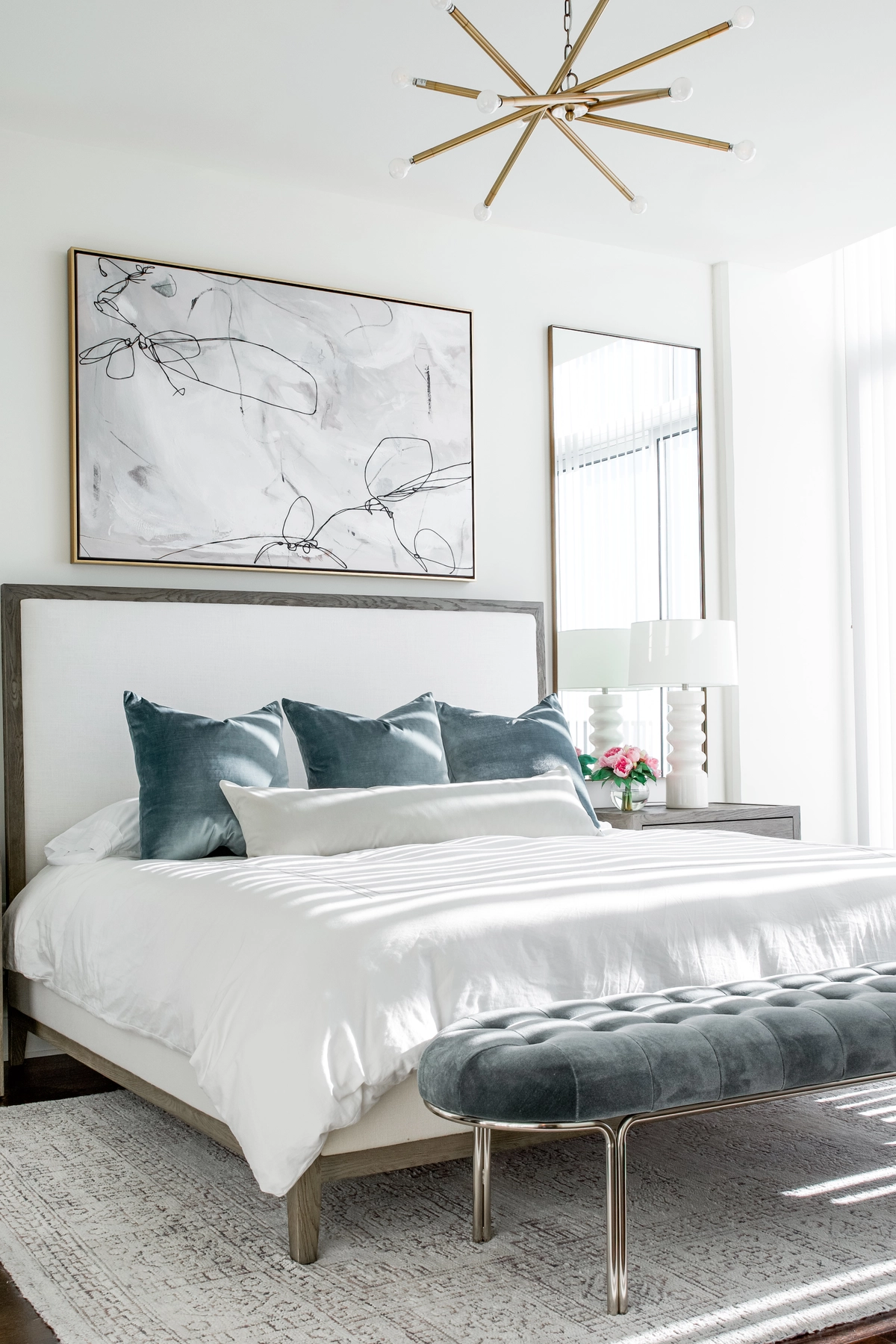 Primary bedroom with a wood-framed upholstered bed, teal velvet pillows, tufted teal bench, abstract art above the headboard, brass sputnik light, and a stacked white lamp on a dark nightstand