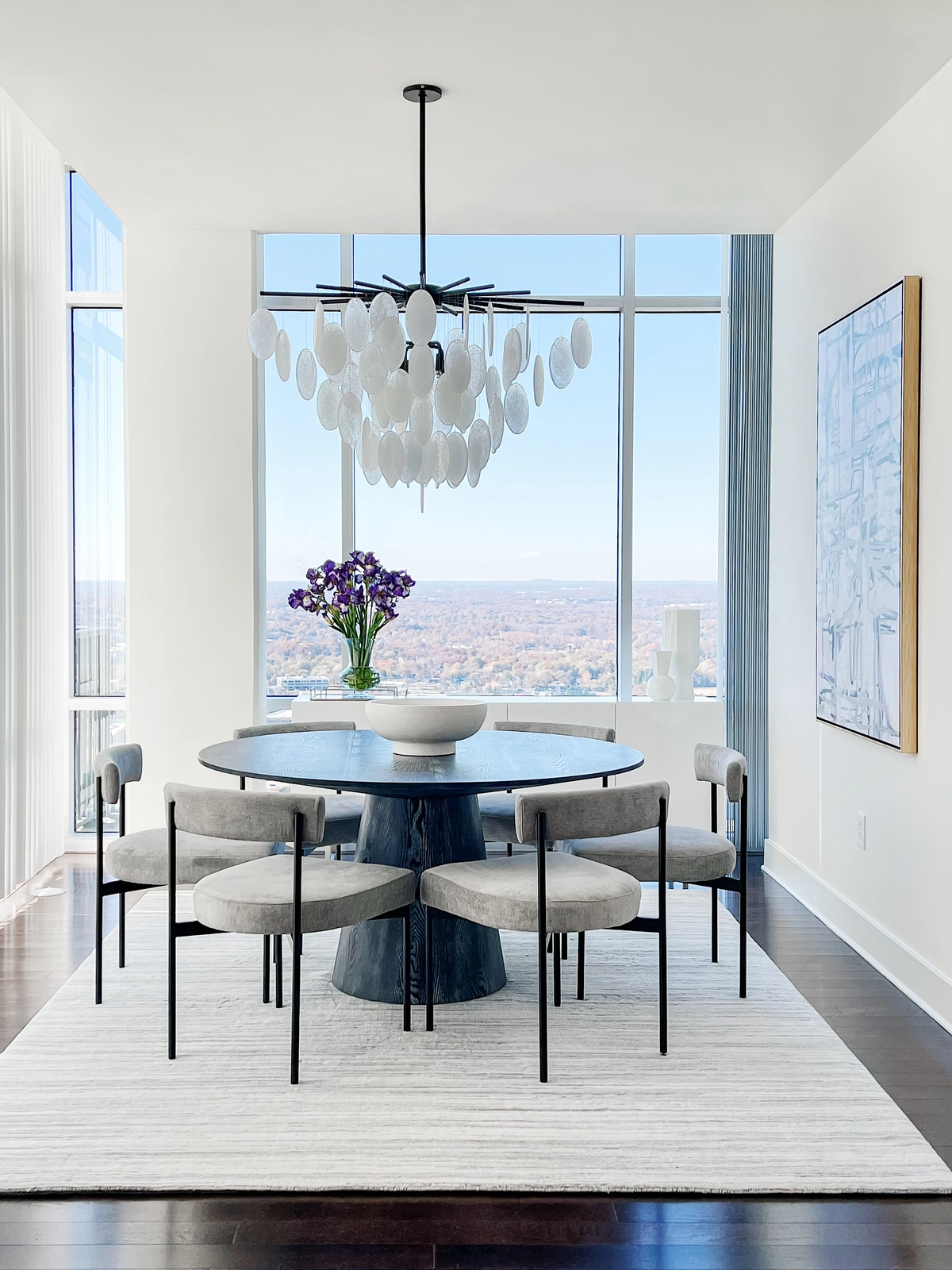 Dining room with a dark round pedestal table, gray velvet dining chairs, a cascading white disc chandelier, purple irises, and corner windows with sweeping city views