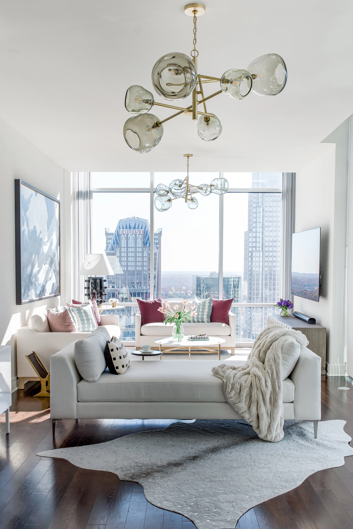 Living room with a smoke glass sputnik chandelier, white sofas with colorful pillows, marble coffee table, fur throw, cowhide rug, and floor-to-ceiling windows with Charlotte skyline views