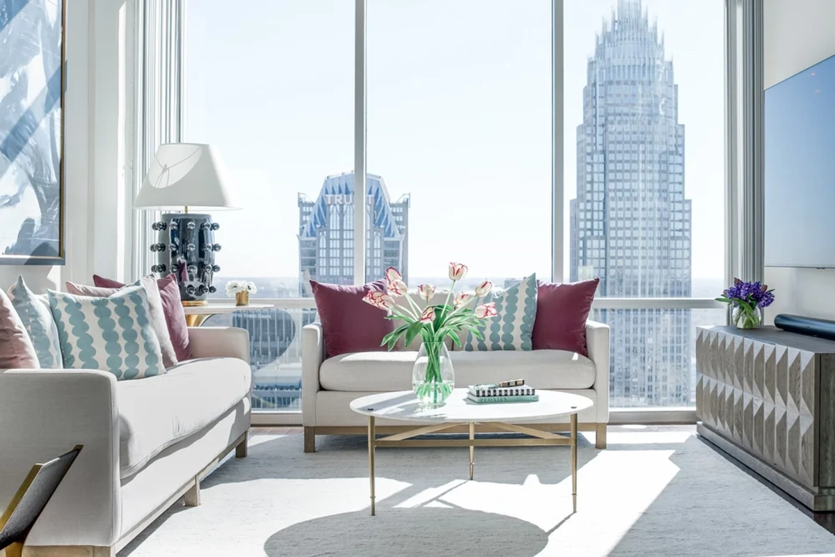 Bright living room with a white sofa, colorful accent pillows in teal and pink, a glass-topped gold coffee table, fresh tulips, and floor-to-ceiling windows framing the Charlotte skyline