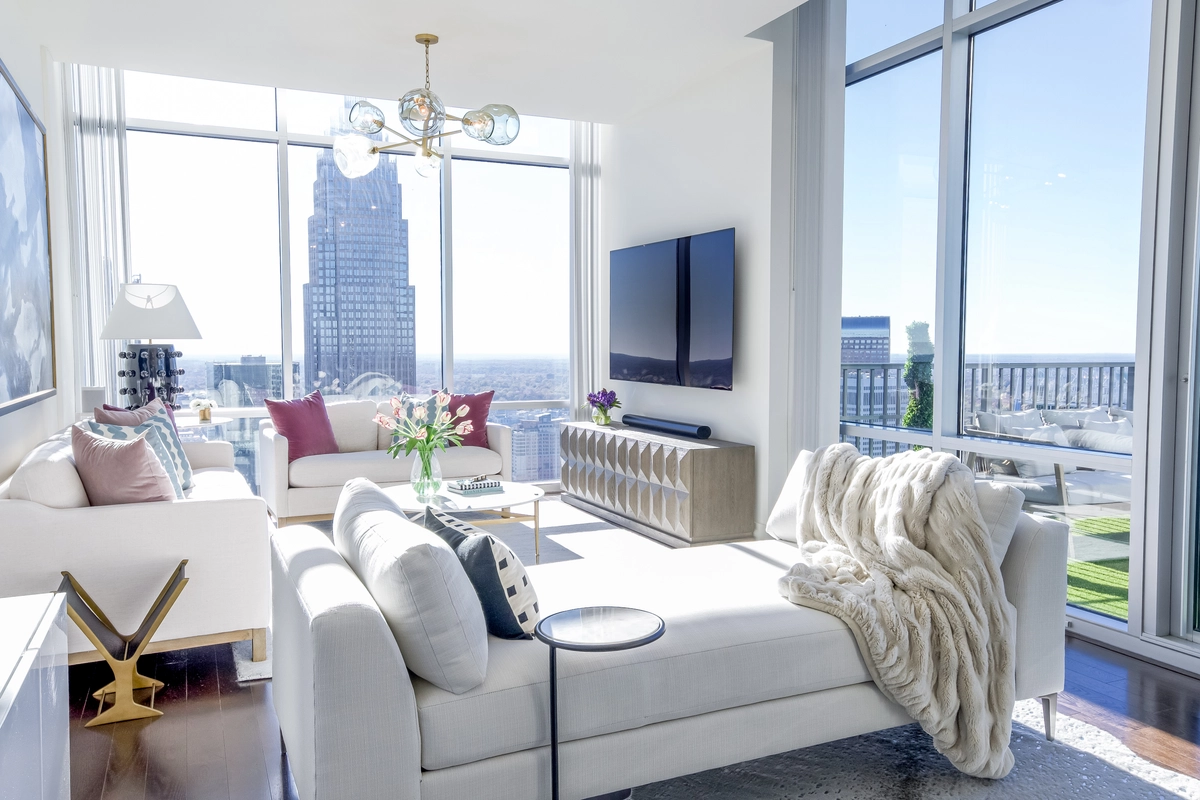 Living room from the sofa looking toward a wall-mounted TV on a tufted credenza, white sofas with accent pillows, sputnik chandelier, and panoramic skyline views through floor-to-ceiling windows
