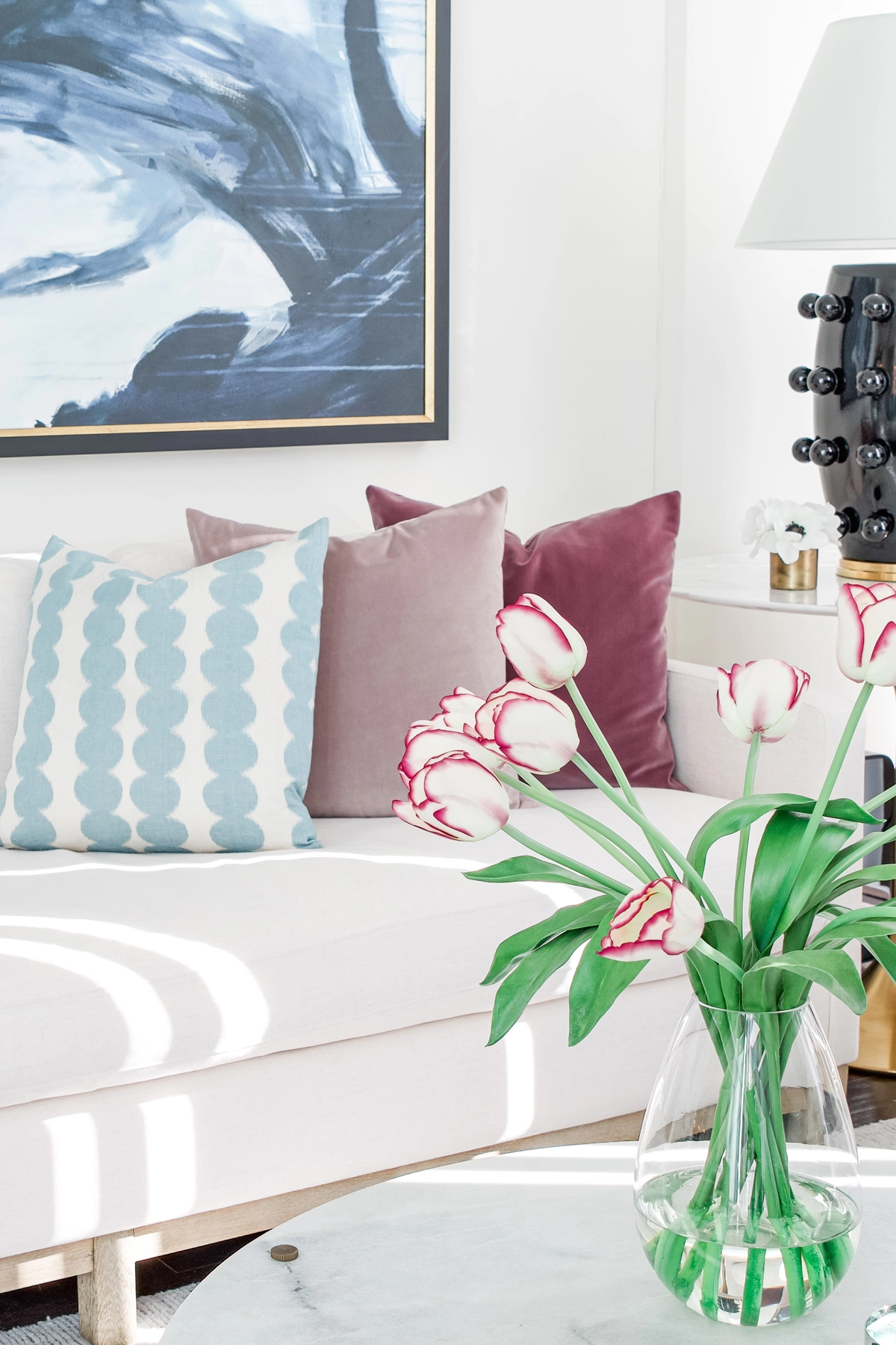Close-up of a white sofa with teal scallop, blush, and burgundy velvet accent pillows, pink-tipped tulips in a glass vase, and a black bobbin table lamp
