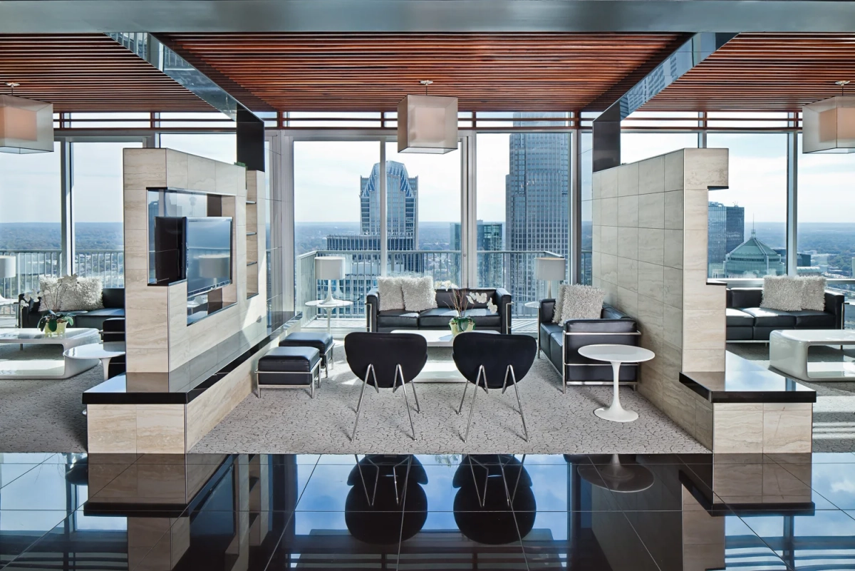 Panoramic sky lounge view from behind the bar counter with black swan chairs, leather seating, a wall-mounted TV on travertine panels, wood-slat ceiling, and sweeping city skyline views