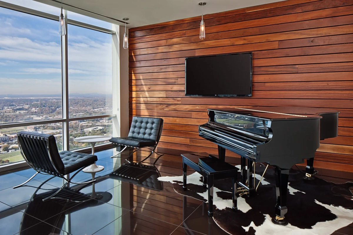Piano lounge with a black grand piano on a cowhide rug, black leather Barcelona chairs, a horizontal wood plank accent wall with a mounted TV, and floor-to-ceiling windows overlooking the city