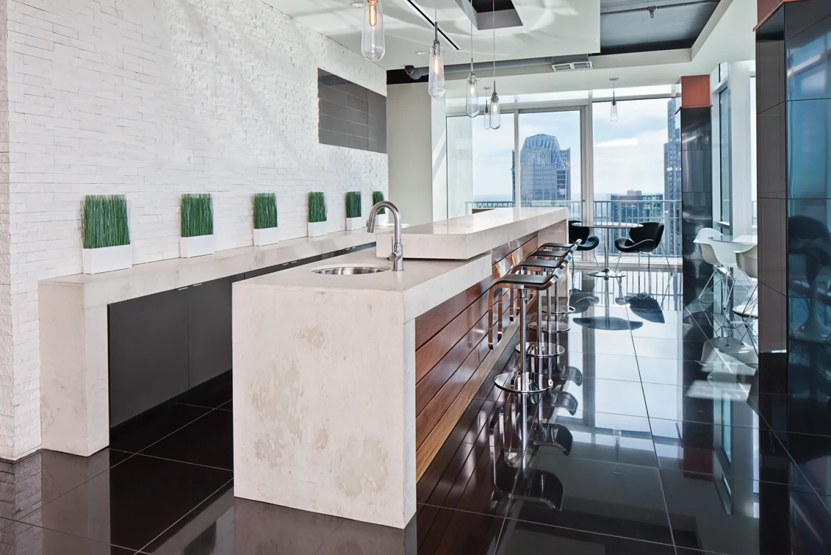 Sky lounge bar with a white marble waterfall-edge countertop, dark wood base, chrome bar stools, glass pendant lights, a white textured stone accent wall with grass planters, and city views