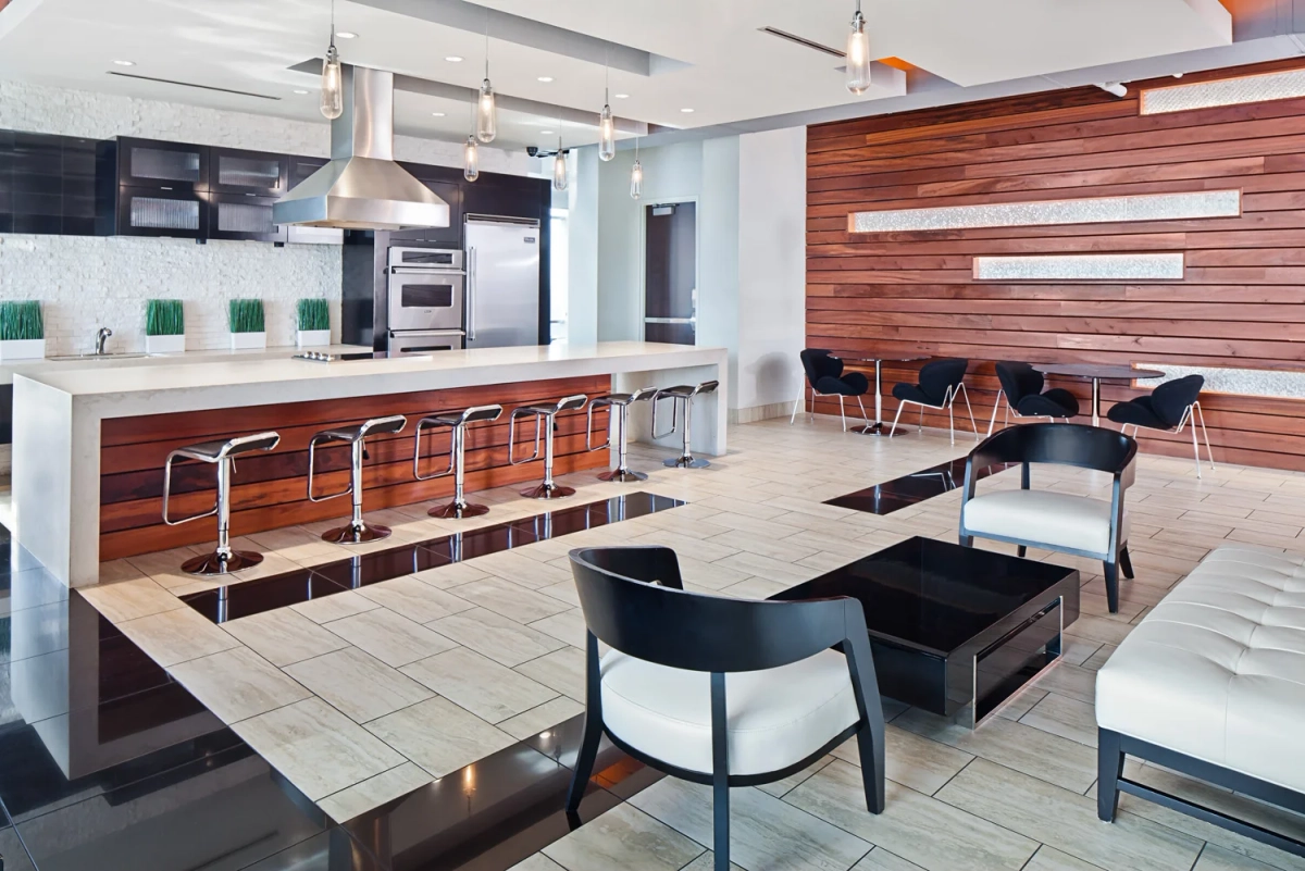 Open-plan sky lounge kitchen and seating area with a long white countertop bar, chrome adjustable stools, a walnut wood accent wall, stainless steel appliances, and industrial pendant lights