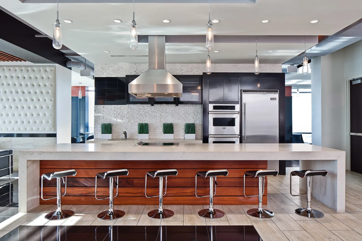 Sky lounge demonstration kitchen with a large white stone island, chrome bar stools with wood accents, stainless steel hood and appliances, glass pendant lights, and a tufted white accent wall